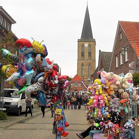 Das Bild zeigt Menschen auf der Kirmes vor dem Kirchturm Emsbüren und einen Luftballon- und Teddy-Stand