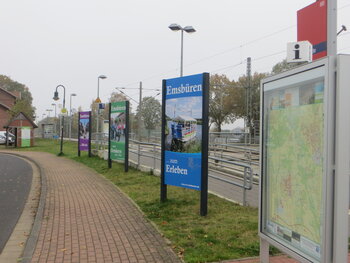 Das Bild zeigt den Bahnhof im Ortsteil Leschede mit touristischen Großformatschildern und dem Ortsplan Emsbüren als Kulisse